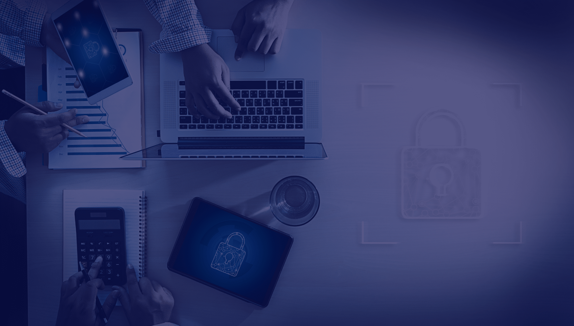 Cyber Security Overhead view of a work desk with a laptop, tablet, calculator, and documents. Hands are typing and writing. A lock symbol overlays the image, suggesting security.